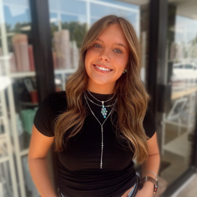 A young woman with long, wavy light brown hair smiles at the camera. She is wearing a black t-shirt and layered silver necklaces. The background shows glass windows and shelves with various items.
