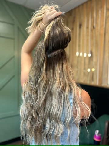 A woman with long, wavy blonde hair lifts the top section of her hair to reveal hair extensions underneath in a salon setting.