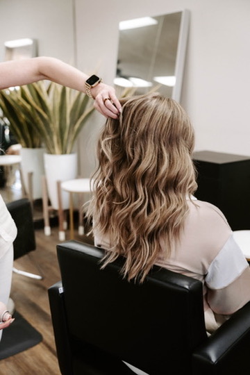 A person with long, wavy, light brown hair sits in a salon chair while a stylist adjusts their hair. Modern decor and large plants are visible in the background.