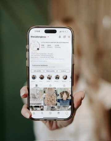 A person holds a smartphone displaying an Instagram profile for a hair salon, featuring a profile photo, bio, and several hair styling images in the feed. The background is blurred.