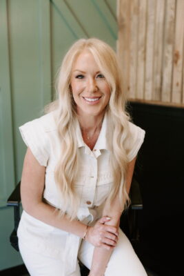 Salon Gloss A smiling woman with long, wavy blonde hair sits on a chair. She is wearing a short-sleeved, white button-up top and white pants. The background features pale green and wooden panel walls.