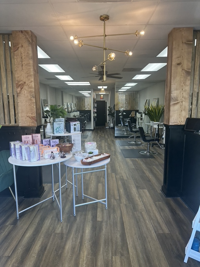 A modern hair salon with wooden floors, product displays on round tables at the entrance, salon chairs and mirrors along both sides, and bright ceiling lights. Tall wooden columns frame the space.