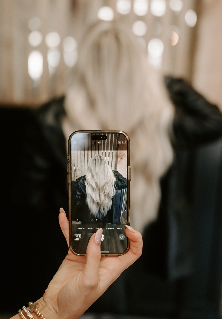 A person holds a smartphone showing a photo of someone with long, wavy, platinum blonde hair and a black jacket, while the actual person sits blurred in the background.