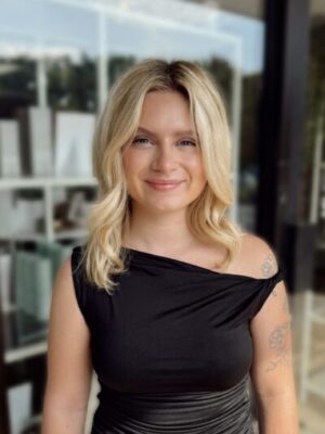 Edited in Tezza with: Vintage & Brightness A woman with blonde hair and a black, off-the-shoulder dress smiles in front of large windows. She has visible tattoos on her arm.
