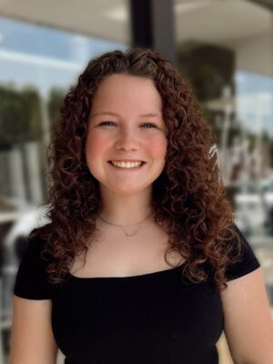 Edited in Tezza with: Vintage A young woman with curly brown hair smiles at the camera, wearing a black top and a delicate necklace, standing outdoors with a blurred background of glass and greenery.