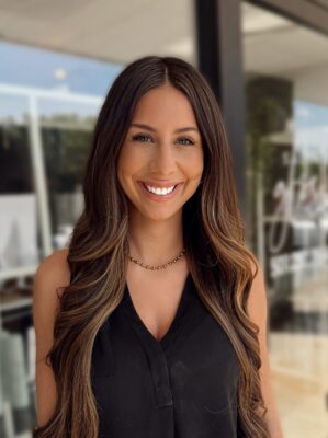Edited in Tezza with: Vintage A woman with long, wavy brown hair and a black sleeveless top smiles outdoors near a glass storefront on a sunny day.
