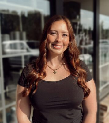 Edited in Tezza with: Vintage & Brightness A woman with long, wavy red hair and a black top stands smiling outdoors in front of a glass window reflecting cars and trees.